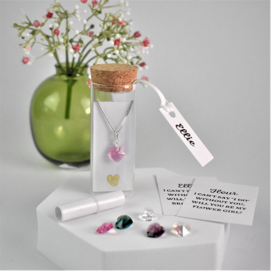 Small glass bottle holds a crystal heart pendant on silver chain. Behind the pendant is a scroll with recipients name and a request for them to be a bridesmaid, flower girl etc. Bottle is finished with a tag bearing the bridesmaid's name.