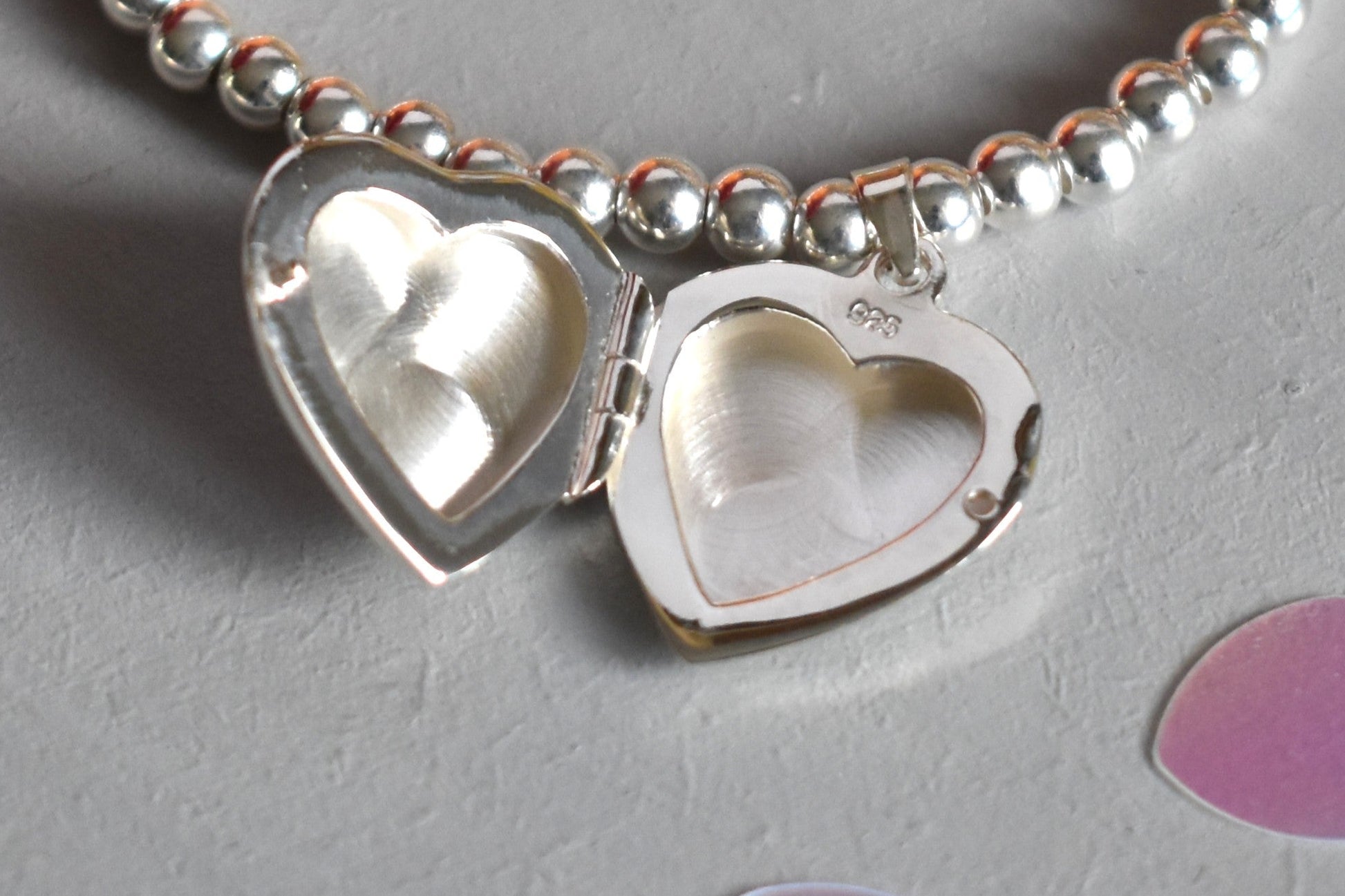 16mm heart shaped sterling silver locket suspended from 4mm sterling silver beads threaded onto strong elastic. The locket opens for you to add two photos of your choice. Shown open.
