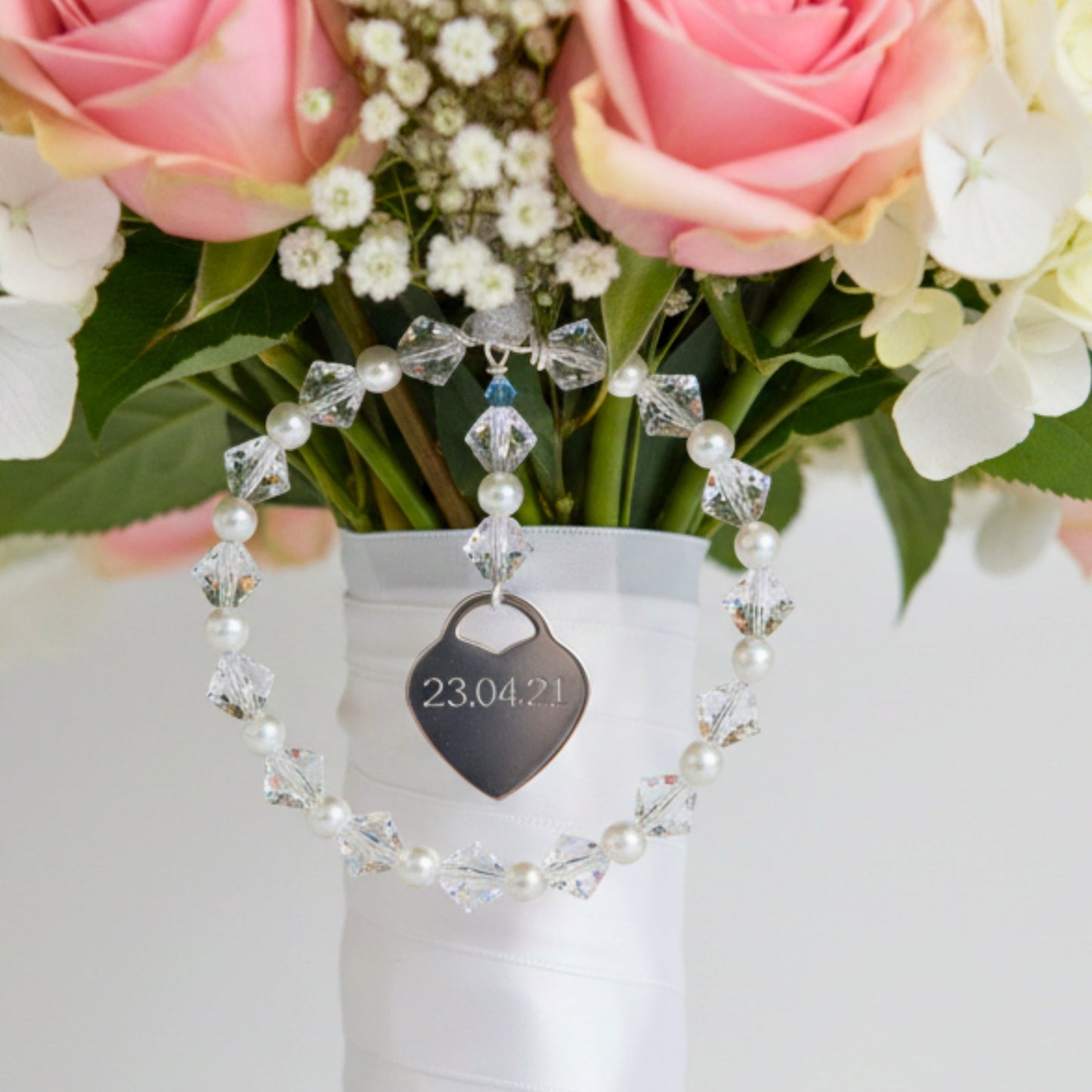 A wedding bouquet charm consisting of a circle of high quality crystals and glass pearls surround a sterling silver heart that can be engraved with names, dates or initials of your choice. Finished with white organza ribbon, the charm can be securely fastened to your wedding bouquet. The tiny blue bead is a nod to the "something blue" tradition.