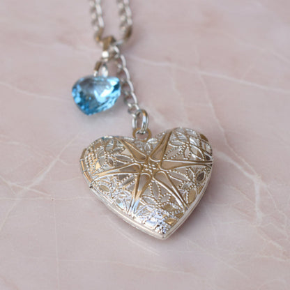 Wedding bouquet locket. A filigree locket that opens to take one photo is suspended from a sterling silver chain and next to a high quality pale blue crystal heart. The chain ends in a lobster clasp so that the charm can be fastened securely to th Bride's bouquet