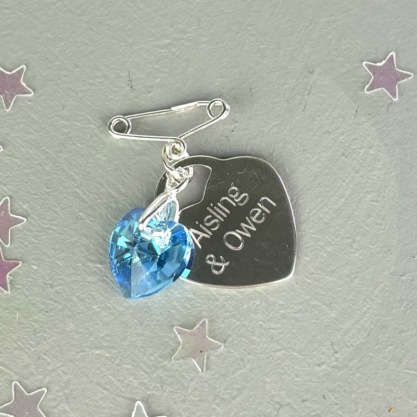 Blue crystal heart suspended from a sterling silver bail and a tiny sterling silver pin. Shown with a sterling silver heart engraved with the names of the couple.
