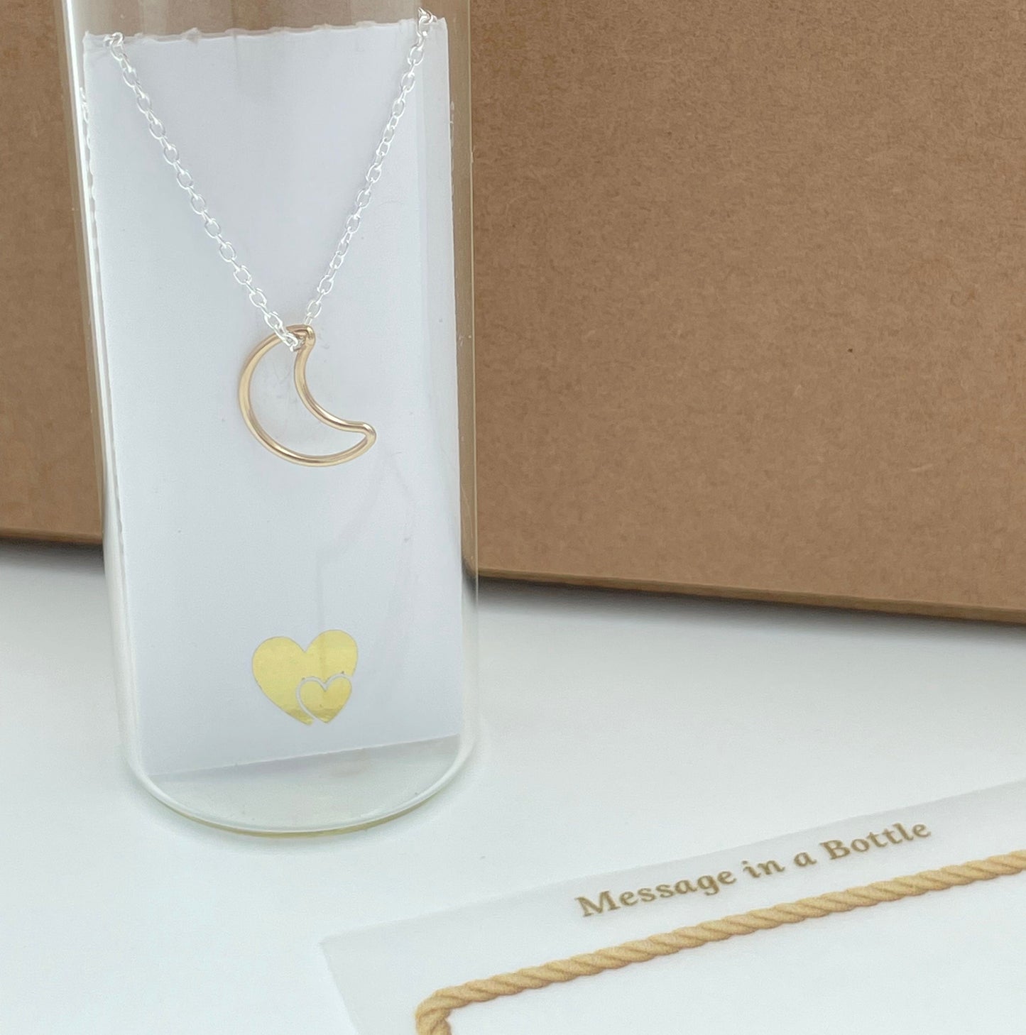 Gold vermeil hollow crescent moon hung from a sterling silver chain and placed in a glass bottle. Behind the card is a parchment scroll for you  to add your own message.