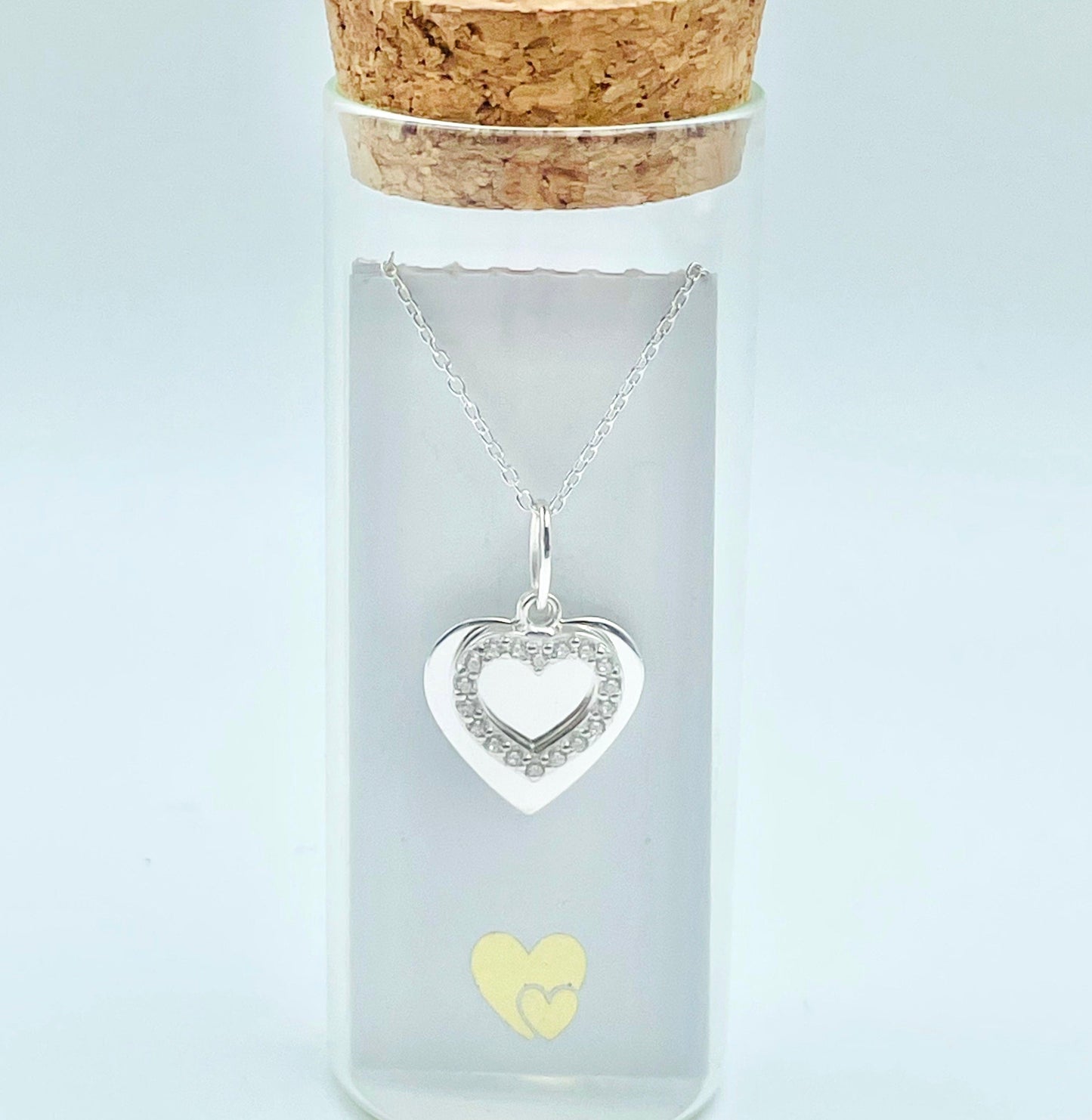 two hearts, one sterling silver and the other an open heart of cubic zircona hung from a sterling slver chain and placed in a glass bottle