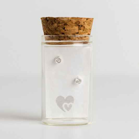 Tiny sterling silver stud earrings with crystal birthstone in a glass bottle with cork lid. Behind the earrings is a parchment scroll for you to add your own message
